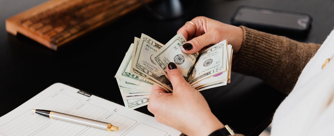 Unrecognizable elegant female in sweater counting dollar bills while sitting at wooden table with planner and pen